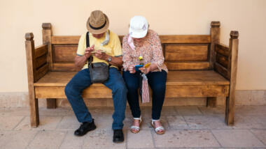 Zwei ältere Menschen sitzen auf einer Holzbank eng nebeneinander. Bei schauen nach unten in ihre Smartphone, sodass man die Gesichter nicht sieht, sondern nur die Kopfbedeckungen.