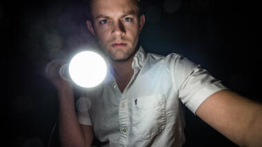 Ein weißer Mann mit weißem Hemd hat eine Taschenlampe in der Hand und leuchtet in das Dunkel.