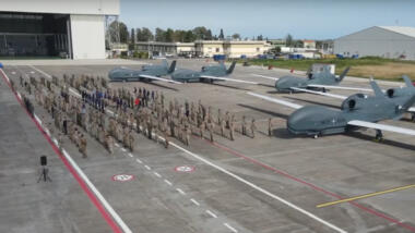 Ein Rollfeld mit 5 Global Hawk, davor stehen mehrere Dutzend Soldat:innen stramm, im Hintergrund ein Hangar.