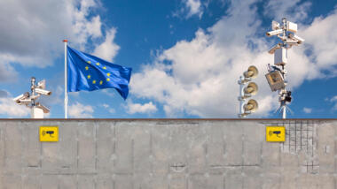Mauer über der eine EU-Fahne weht, links und rechts davon Überwaxchungskameras.
